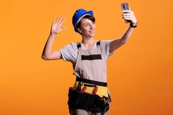 Happy female builder talking on online video call with smartphone over yellow background, using videoconference call. Cheerful constructor chatting on remote teleconference on mobile device.
