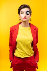A young woman with a pirate patch over one eye. A sly expression on face. Short haircut, red jacket, yellow T-shirt, bright yellow background in the studio, earrings. A modern pirate boss girl