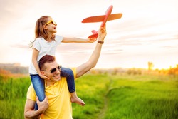 Cheerful man smiling and carrying excited girl on shoulders while playing with red aircraft together against cloudy sky on sunny summer day