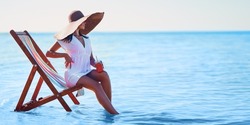  Beautiful caucasian girl in sunhat and white tunic sitting on the beach chair at seaside while holds cocktail. Loking in sideways. Summer vacation. Skin care