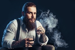Portrait of handsome elegant brutal bearded man with cigar and a glass whiskey in a dark room. Tattoos hands 