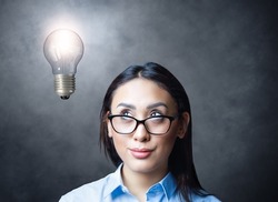 Pensive young woman with eyeglasses looking up to lightbulb light on concrete wall background. Creative thinking concept.
