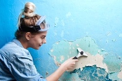 Girl with scraper in hand peels off a layer of cracked blue paint on the wall in the apartment, woman makes repairs