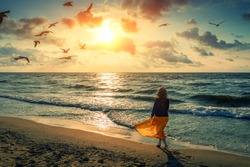 Seascape at sunrise with beautiful sky. Woman on the beach. Young happy woman in a yellow fluttering dress walks along the seashore. The girl looks at the magical sunrise. Seagulls fly over the beach