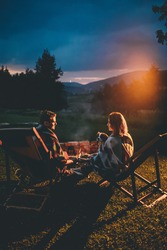 Romantic Couple With Cute Dog Relaxing In Campsite With Fire Pit. Burning Campfire with mountain landscape with night sky over the forest and hills. Getaway in wild nature.