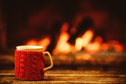 Cup of hot drink in front of warm fireplace. Holiday Christmas concept. Mug in red knitted mitten standing near fireside. Cozy relaxed magical atmosphere in a chalet.