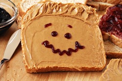 Making peanut butter sandwiches with personality!  Fun smiley face drawn on with jam. Creamy peanut butter with jam on whole grain wheat bread on wood cutting board. Macro with shallow dof.