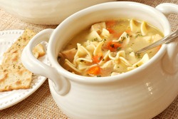 Chicken noodle soup in cream colored ceramic bowl with handles.  Plate of crackers and soup tureen in background.  Closeup with shallow dof.