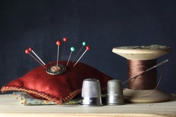 OLD WOODEN COTTON THREAD REEL AND NEEDLE, PIN CUSHION WITH GLASSHEAD STEEL PINS AND THIMBLES ON A WOODEN SURFACE