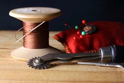 TABLE TOP IMAGE OF A TRACING WHEEL, PIN CUSHION AND COTTON REEL ON A WOODEN SURFACE
