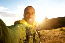 Portrait of happy young black man hiking with backpack taking selfie and pointing to sunset