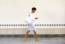 Full length side portrait of a young woman walking and sending text message on cell phone 