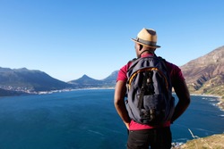 Rear view portrait of young african man on holiday with hat and backpack looking at the sea