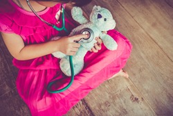 Top view of child playing doctor or nurse with plush toy bear with bright sunlight at home. Happy girl listens a stethoscope to toy. Playful girl role playing. Vintage tone effect.
