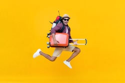 Excited happy young Asian man tourist with luggage jumping isolated on yellow studio background
