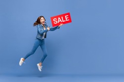 Excited Asian woman in casual jean clothes jumping with red sale sign in hand isolated on light blue background with copy space