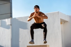 Fit shirtless athletic man jumping up to plyometric wood box outdoors on building rooftop, home workout exercise concept