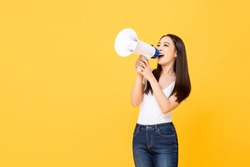 Portrait of smiling cheerful young pretty Asian woman holding megaphone making announcement in isolated studio yellow background