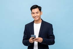 Waist up portrait of Young smiling handsome Asian man in semi formal suit using mobile phone in light blue isolated studio background