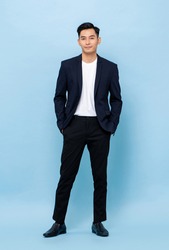 Full length portrait of young handsome Asian businessman standing and posing with hands in pockets on light blue studio background