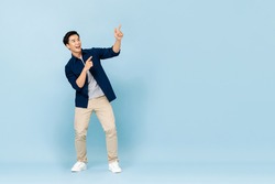 Full length portrait of smiling young handsome Asian man pointing fingers to empty space aside in isolated studio light blue background