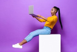 Excited African American woman holding and watching  laptop computer while sitting on stool in isolated purple color studio background