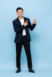 Young handsome smiling Asian man wearing semi-formal suit looking at camera and pointing hands up in blue color studio isolated background