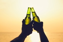 Couple hands holding beer bottles and clanging, celebrating on holiday at the beach in summer sunset