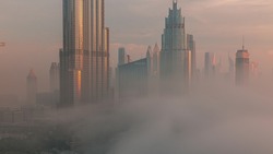 Aerial view of Dubai city early morning during fog timelapse. Sunrise at futuristic city skyline with skyscrapers and towers from above. Sun reflected from glass surface