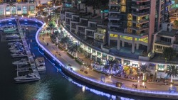 Waterfront promenade with palms and many restaurants in Dubai Marina aerial night timelapse. Yachts and boats floating on water near pier