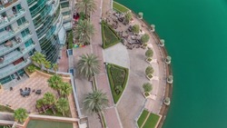Waterfront promenade with palms in Dubai Marina aerial timelapse. Tables and chairs of restaurant near trees. Yachts and boats floating on water