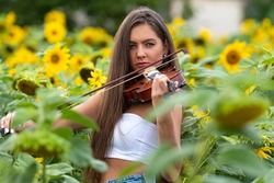young slim brunette with long hair standing in sunflower field and playing violin