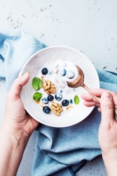Hands holding a healthy breakfast bowl with yoghurt, blueberries and walnuts. Captured from above (top view, flat lay). Pastel blue stone background with free copy (text) space.
