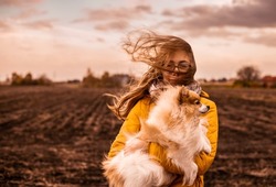 Windy dull autumn day. Outdoor portrait of caucasian teenage girl with a pomeranian dog. Long blond hair blowing in the strong wind - gale. Country fields landscape as background.