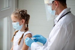 A doctor in a protective suit makes the child  in a medical mask a shoulder shot . Coronavirus vaccination. Covid-19 vaccine. Doctor vaccinating child. Kids at clinic. Little girl getting flu shot. 