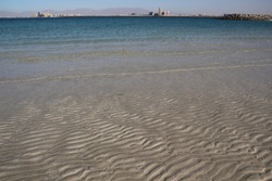 Ripple texture of  white sea coast sand in water with background of sea, buildings, mountains, blue sky