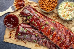 2 racks of ribs sitting on brown paper with barbeque sauce brush, sides of cole slaw, baked beans and garnished with fresh thyme herbs