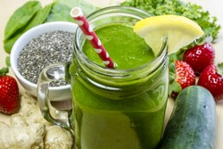 Healthy green juice smoothie surrounded by whole fruits, vegetables and chia seeds with lemon garnish and red polka dot straw