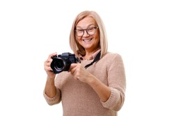 Portrait of smiling senior woman photographer 60-65 years old with blonde hair standing, holding photocamera in hands and looking at camera. Concept of lifestyle. Isolated on white. 