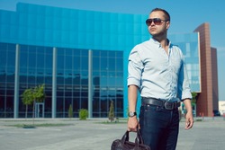 Men Shopping concept. Portrait of attractive proud man in trendy casual clothing with leather bag and sunglasses posing over shopping mall. Sunny spring weather with blue sky. Outdoor shot