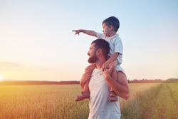 Small boy is sitting on his father??s shoulders and pointing at the sun in the field during beautiful sunset 