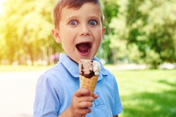 Small boy with charming blue eyes is holding ice-cream and making a surprised face while walking in the park 