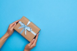 Top view of hands placing a wrapped box, tied with a ribbon. Gift box lying on blue background 