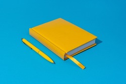 Photo of closed yellow notebook and ball-point pen over blue background. Minimalist image of closed diary and yellow pen as back to school concept.
