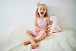 Adorable little girl in pink dress and golden crown dressed as princess playing with unicorn. Kid having fun with soft toy. Child playing with stuffed toy