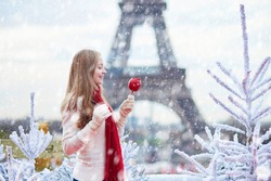 Girl with caramel apple on a Parisian Christmas market during snowfall near white snowy Christmas trees and with the Eiffel tower in the background