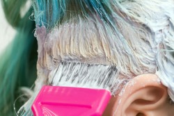 Close-up of hairdresser using pink brush while applying paint to female customer with emerald hair color during process of bleaching hair roots in hair salon. Barber point of view shot