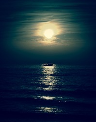 Silhouette of the fisherman or leisure boat sailing toward the moon. Dark sky and clouds. Moonwalk. Reflection in water. Beautiful seascape in the night. Harmony with nature idea. Mystery background.