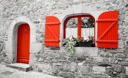 Old stone house with red wooden shutters and red door. Boxes with red and white flowers on the window. Brittany, France. Retro aged photo. 