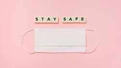 stay safe, plastic letter tiles, and white fabric face mask on pink paper background, flat lay